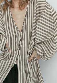 Pasiasta, oversize bluzka w kolorze ecru i brązowym. Posiada głęboki dekolt w serek oraz zapięcie na guziki. Wykonana z lekkiego materiału z szerokimi rękawami.