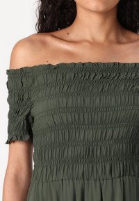 Off-shoulder klänning i olivgrön tyg, med en smockad byst och korta volangärmar med en mjuk textur.