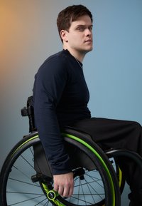 Egy fiatal férfi fekete kerekesszékben ül, zöld díszítéssel, és sötétkék, hosszúujjú pólót visel. A háttér kék és narancs színátmenete jellemzi.