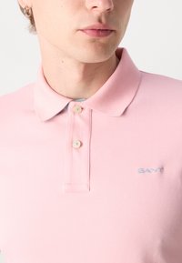 Trumpas rožinis polo marškinėliai su klasikiniu apykakle, turintis dvi sagas, šviesiai mėlyna logotipo siuvinėjimas krūtinėje ir tekstūruota medžiaga.