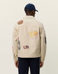 Μπεζ μπουφάν με διάφορα εκτυπωμένα γραφικά στην πλάτη, συμπεριλαμβανομένων φρούτων, ενός μπουκαλιού και ενός παιχνιδιού. Χαρακτηρίζεται από ύφασμα 100% βαμβακιού και τυπικό σχεδιασμό για γιακά.