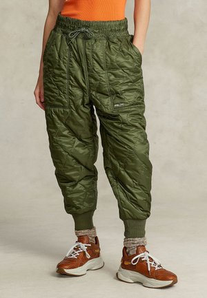 Woman wearing olive green quilted jogger pants with elastic cuffs and brown sneakers, standing with hands in pockets.