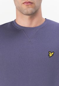 Felpa viola con colletto a coste e tessuto leggermente testurizzato. Presenta un piccolo logo giallo ricamato nell'area sinistra del petto.