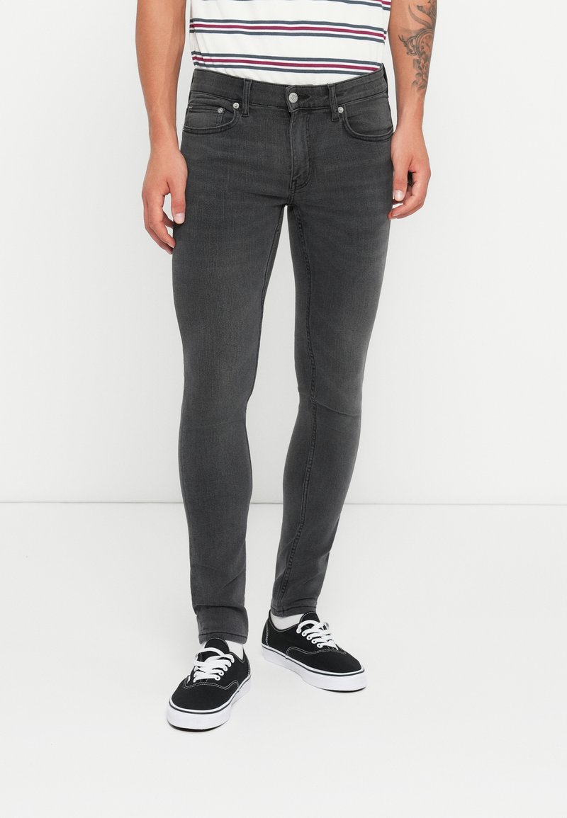 Jeans de mezclilla gris ajustados con cinco bolsillos, cierre de botón de metal y un ligero desgaste. Combinados con zapatillas negras y una camisa a rayas.