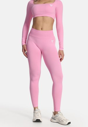 Frau trägt passendes rosa langärmeliges Crop-Top und Leggings mit weißen Sportschuhen und steht vor weißem Hintergrund.