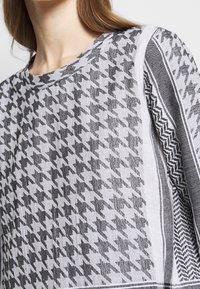 Femme portant un haut à motif pied-de-poule noir et blanc avec des rayures sur les manches et un col rond.