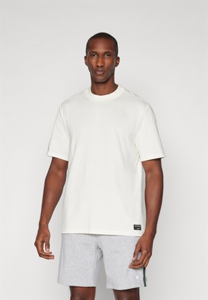 White short-sleeve t-shirt with a classic crew neck, made of smooth cotton, featuring a small logo tag near the hem. Worn with grey shorts.