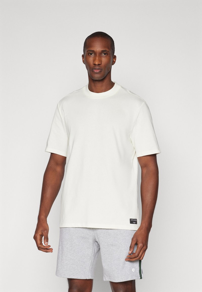 Vit kortärmad t-shirt med klassisk rund halsringning, gjord av mjuk bomull, med en liten logotag nära fållen. Bärs med grå shorts.