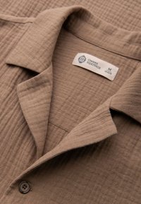 Chemise en tissu brun texturé avec un col plié, un bouton visible et une étiquette indiquant Ombre Heritage taille moyenne.