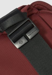 Detail van een kastanjebruine tas met gestructureerde stof, zwarte riem en een zwarte metalen gesp met gegraveerd logo. Het design combineert duurzaamheid en functionaliteit.