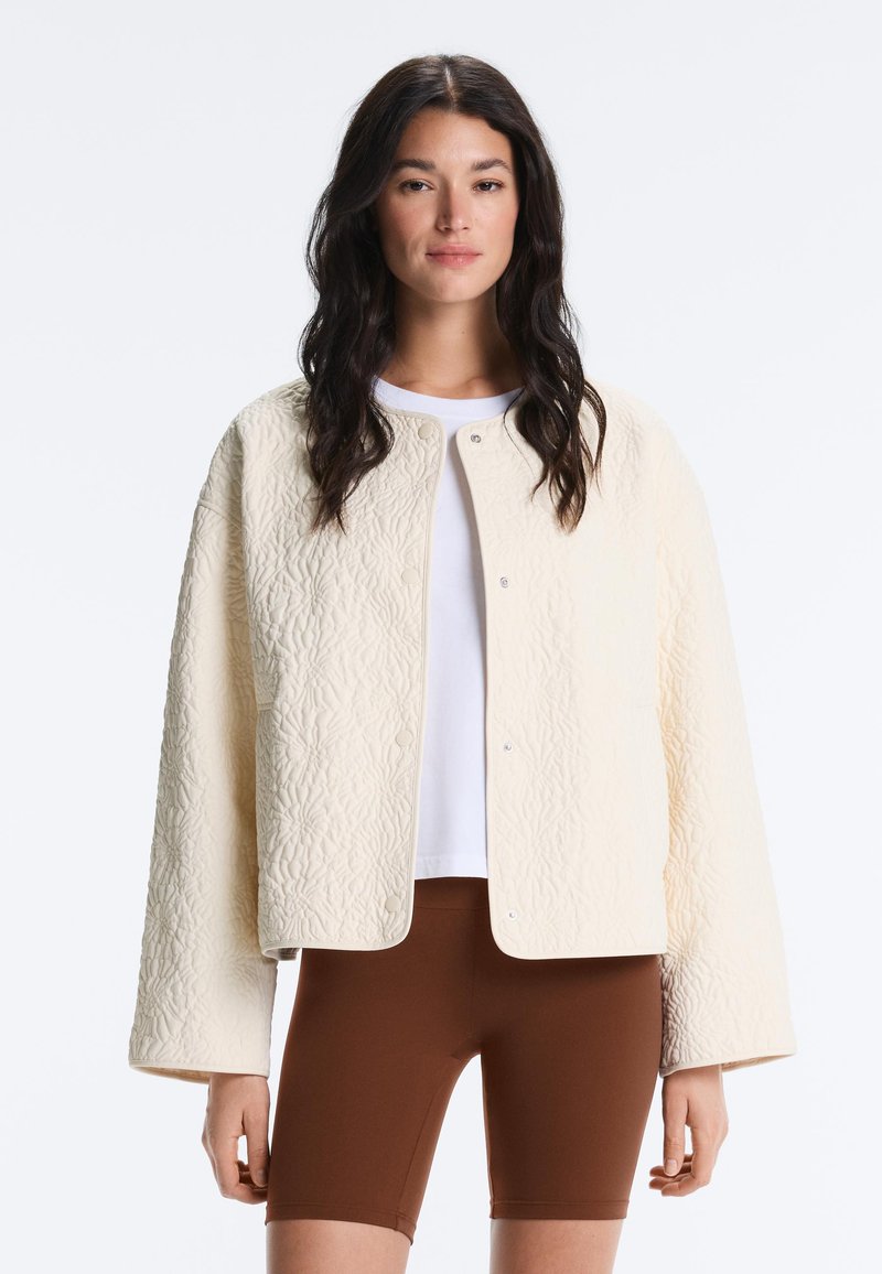 Femme aux longs cheveux foncés portant une veste matelassée crème texturée par-dessus une chemise blanche et un short marron ajusté, debout devant un fond blanc.