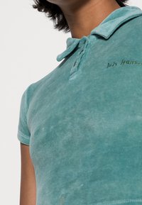 Tyrkysové polo tričko vyrobené z měkké tkaniny, s límečkem, dvěma knoflíky a vyšitým detailem nad levou hrudí. Hladká textura.