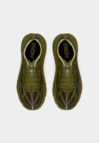 Ζευγάρι αθλητικών παπουτσιών HOKA σε ελιά πράσινο χρώμα με δέσιμο κορδονιών και μαύρες εσωτερικές σόλες, εμφανίζονται από πάνω σε απλό λευκό φόντο.