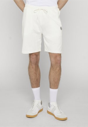 Mann iført hvite shorts med snøring, hvite crew-sokker og hvite joggesko med gummisåler, stående mot en enkel lys bakgrunn.