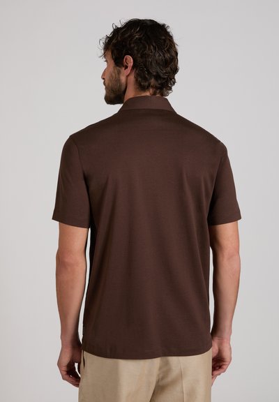 Mann mit lockigem Haar, der ein einfarbiges braunes Poloshirt mit kurzen Ärmeln und beige Hosen trägt, von hinten vor einem hellgrauen Hintergrund gezeigt.