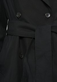 Melns tranšejmētelis ar gludu audumu, dubultpogājuma dizainu un pašpiesaistes jostu. Aprīkots ar apaļām melnām pogām aiztaisīšanai.