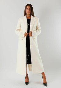 Lungo cappotto crema realizzato in morbido tessuto texturizzato con una struttura tailored, dotato di colletto a reverse e tasche frontali, abbinato a un vestito nero e tacchi.