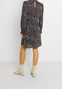 Jurk met lange mouwen met een paisleyprint in donkerblauw, bruin en wit, voorzien van een keyhole-detail aan de achterkant. Gecombineerd met crèmekleurige laarzen met blokhak.