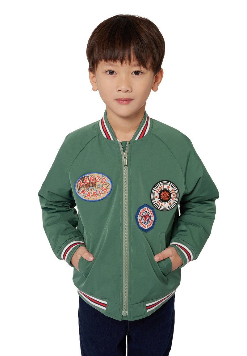 Veste bomber verte avec col et poignets côtelés rayés, ornée de plusieurs patches brodés. Fermeture éclair à l'avant et poches latérales.