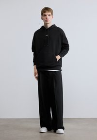 Μαύρο φούτερ με κουκούλα και μπροστινή τσέπη με κορδόνι, μαύρο παντελόνι φαρδύ, και άσπρα αθλητικά παπούτσια, όλα σε μια λεία, μίνιμαλ σχεδίαση.