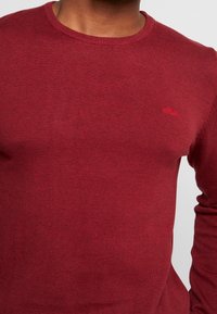 Rød striktrøje med rund halsudskæring, lange ærmer og et diskret logo broderet i det øverste venstre brystområde. Blød tekstur.
