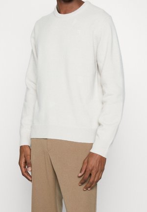 Mann trägt einen schlichten weißen Langarmpullover und beige Hosen, steht vor einem hellen Hintergrund.