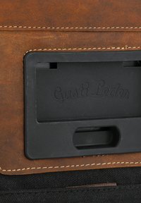 Pelle marrone con cuciture bianche a contrasto e un compartimento in plastica nera etichettato "Gusti Leder". Superficie texture con una finitura liscia.