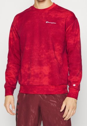 Man die een rood tie-dye Champion sweatshirt met lange mouwen en bijpassende rode broek draagt.