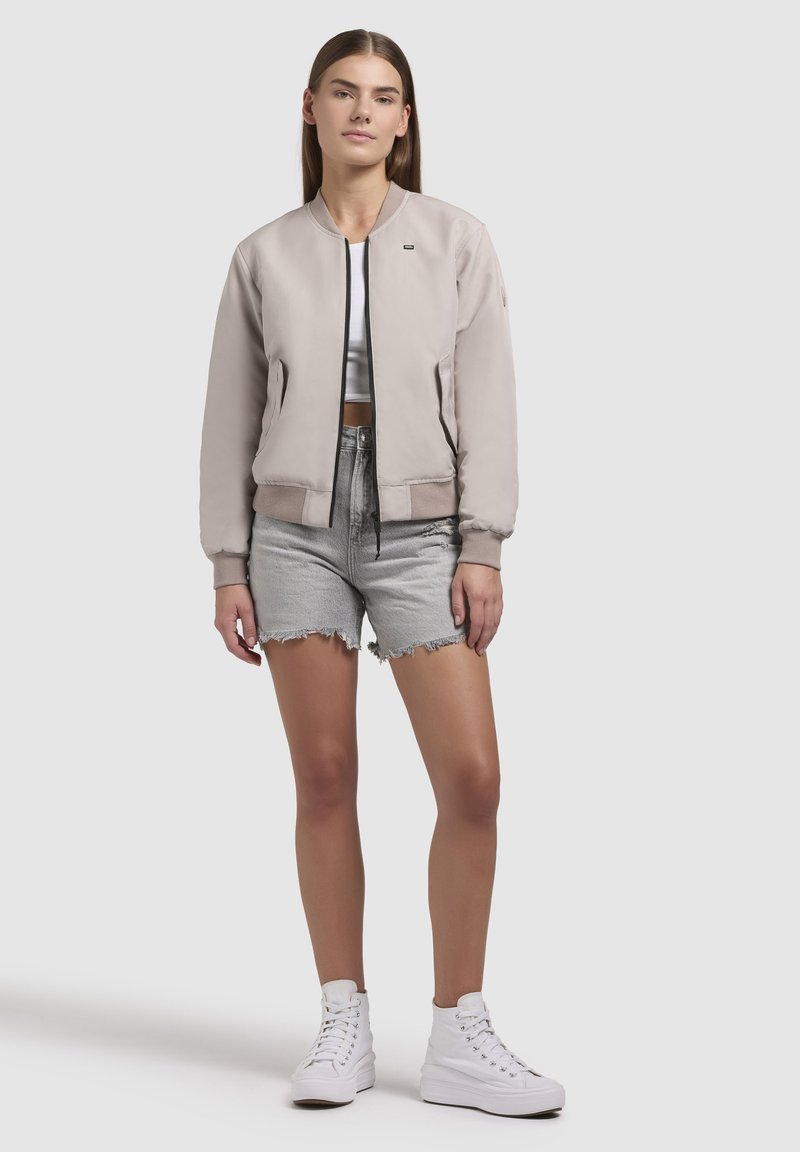 Helebeež bomber-jakk ribatud mansettide, musta tõmbluku ja kahe ees oleva taskuga, kombineerituna hallide teksashortside ja valgete kõrgete tennisetossudega.