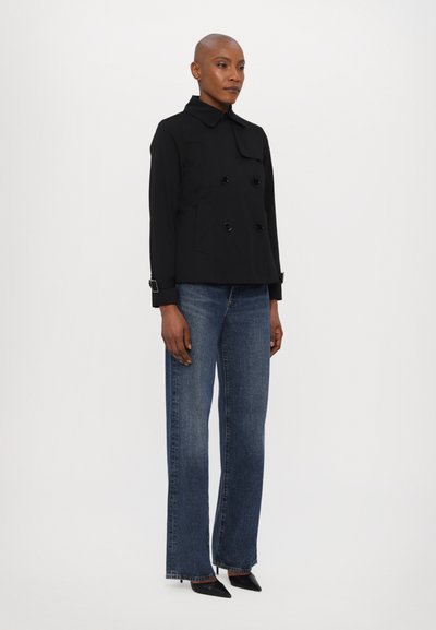 Veste noire à double boutonnage avec un col structuré, ornée de grandes boutons noirs et de poches latérales, associée à un jean bleu taille haute.