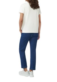 Witte T-shirt met korte mouwen in combinatie met donkerblauwe jeans en witte sneakers. De stof lijkt glad; de jeans hebben een rechte snit.