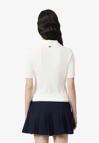 Polo shirt blanc à manches courtes avec des motifs verticaux texturés et le logo Lacoste dans le dos. Associé à une jupe plissée bleu marine.