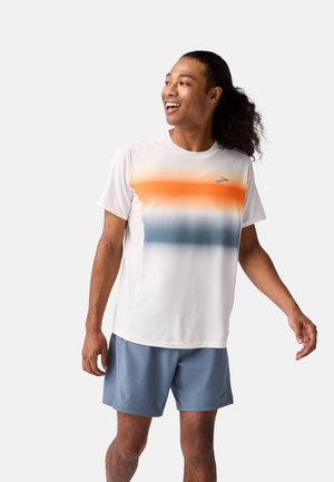 Mann mit langen lockigen Haaren, der ein weißes T-Shirt mit orange- und blauen Streifen und blaue Shorts trägt, lächelt und schaut zur Seite.