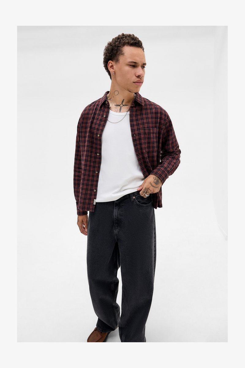 Jovem com tatuagens, vestindo uma camisa de xadrez vermelha sobre uma t-shirt branca, calças jeans pretas e sapatos castanhos, de pé com a mão no bolso.