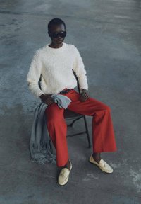 Weißer strukturierter Pullover, rote weite Hosen, grauer Schal, der über den Schoß gelegt ist, und hellgelbe Loafers mit Quasten. Schwarze Sonnenbrille rundet den Look ab.