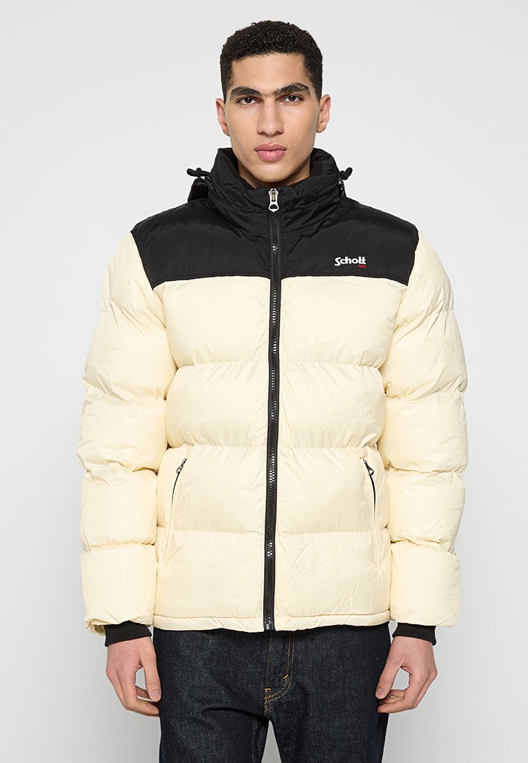 Junger Mann in einer cremefarbenen und schwarzen Steppjacke mit Frontreißverschluss und Reißverschlusstaschen, der vor einem schlichten hellen Hintergrund steht.