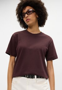 Karmīna kokvilnas t-krekls ar īsām piedurknēm, apaļu dekoltē un brīvu piegriezumu, apvienots ar melnu jostu ar sudraba akcentiem un baltām biksēm.