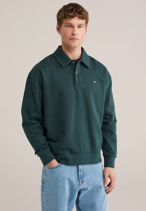 Jonge man die een donkergroene poloshirt met lange mouwen en lichtblauwe spijkerbroek draagt, staand met één hand in de zak tegen een effen achtergrond.