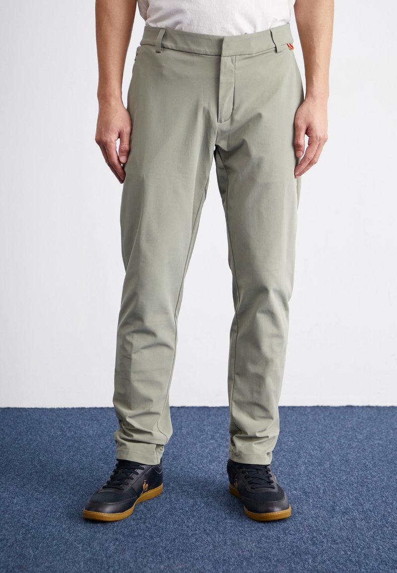 Hombre con pantalones ajustados color gris claro y zapatillas negras con suelas de goma, de pie sobre una alfombra azul.