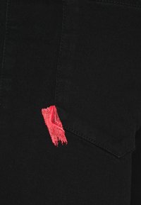 Tela de mezclilla negra con un bolsillo visible que presenta un acento deshilachado de color rojo brillante. La textura es suave, mostrando detalles de costura limpios.