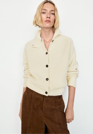 Femme blonde portant un pull côtelé crème boutonné et un pantalon en daim marron, debout sur un fond blanc uni.