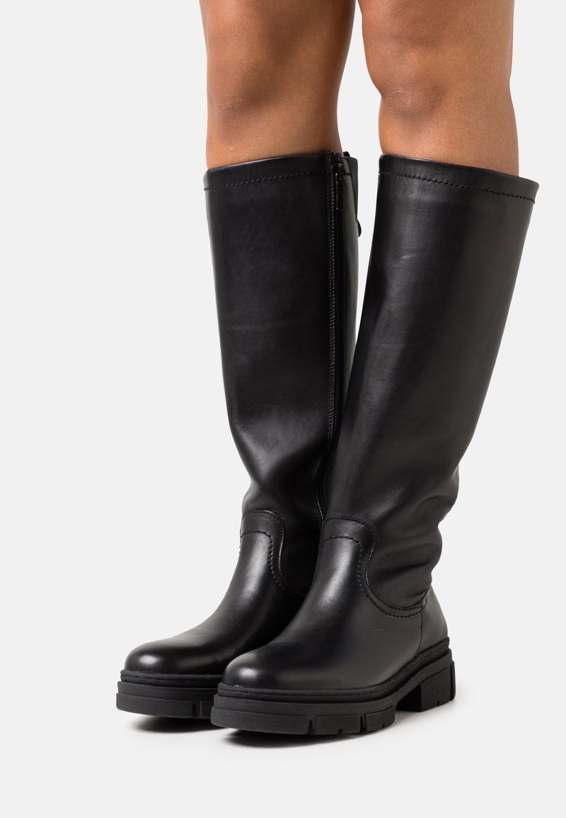 Bottes en cuir noires montant jusqu'aux genoux avec semelles épaisses et fermetures éclair latérales, portées sur des jambes nues sur un fond clair uni.