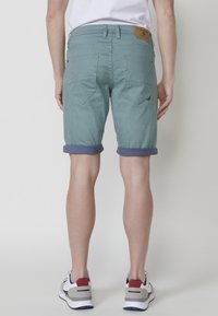 Shorts turquoise clair avec des revers, dotés de poches arrière et d'une étiquette en cuir à la taille, fabriqués dans un tissu léger.