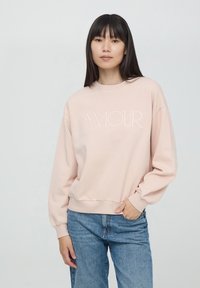 Jasnoróżowy sweter z haftem "AMOUR" na środku, o długich rękawach i luźnym kroju, zestawiony z niebieskimi dżinsami.