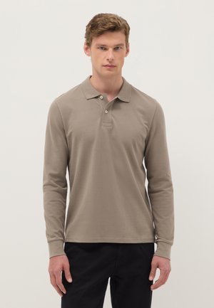 Joven de pie vistiendo una camisa tipo polo beige de manga larga y pantalones negros contra un fondo liso y claro.