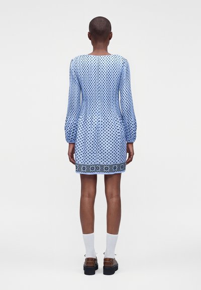 Femme aux cheveux courts portant une robe bleue à manches longues avec un motif losanges noirs et une bordure décorative, chaussettes blanches et chaussures marron, de dos.