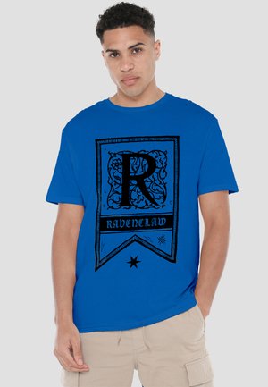 Young man wearing a blue t-shirt with a black "R" and "Ravenclaw" text, paired with light beige cargo shorts.