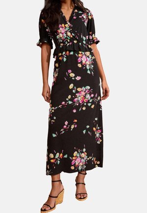 Mujer con vestido cruzado floral negro con mangas abullonadas y detalles de volantes, combinado con sandalias de cuña negras con tiras.