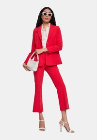 Completo su misura rosso con giacca aderente e pantaloni a campana, abbinato a una camicia bianca. Gli accessori includono una borsa bianca e occhiali da sole a occhio di gatto.