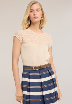 Jeune femme portant un haut beige à manches courtes et une jupe plissée rayée bleue, blanche et marron avec une ceinture marron, debout devant un fond neutre.